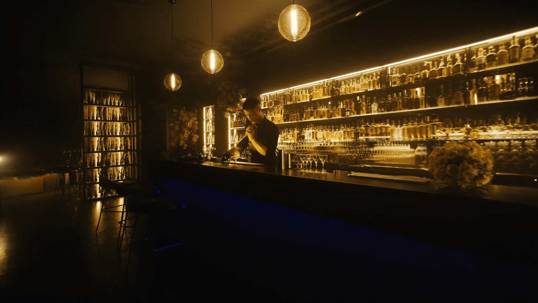 The long NYX bar lit by golden pendant lights, a bartender mixing beneath glowing shelves.